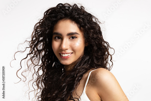 A pretty girl with thick black and curly hair and glowing skin smiles and looks at the camera.