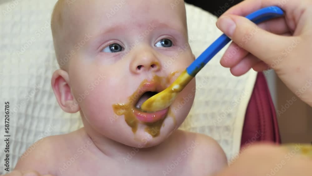 Mom feeding little boy with broccoli puree. Child at the age of six months eats broccoli while sitting on a baby chair. Slow motion shot. Mom feeding little boy with broccoli puree. Child at the age of six months eats broccoli while sitting on a baby chair. Slow motion shot.