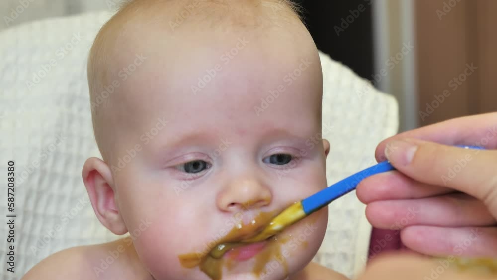 Mom feeding little boy with broccoli puree. Child at the age of six months eats broccoli while sitting on a baby chair. Slow motion shot. Mom feeding little boy with broccoli puree. Child at the age of six months eats broccoli while sitting on a baby chair. Slow motion shot.
