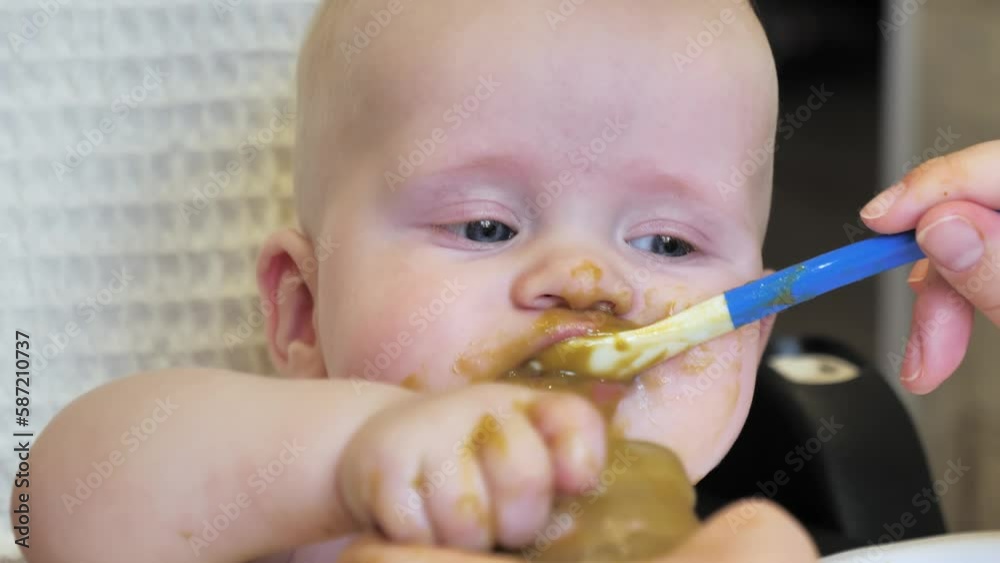 Mom feeding little boy with broccoli puree. Child at the age of six months eats broccoli while sitting on a baby chair. Slow motion shot. Mom feeding little boy with broccoli puree. Child at the age of six months eats broccoli while sitting on a baby chair. Slow motion shot.