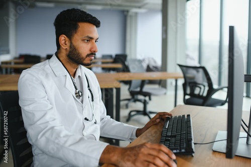 Wallpaper Mural People, occupation and medicine concept. Smiling male Indian Arab doctor in white coat, sitting in medical office at desk with laptop Torontodigital.ca