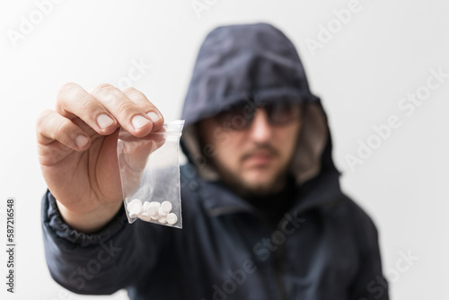 Dangerous man in hooded jacket and sunglasses holding plastic transparent bags with white pills, drug dealer offering narcotics