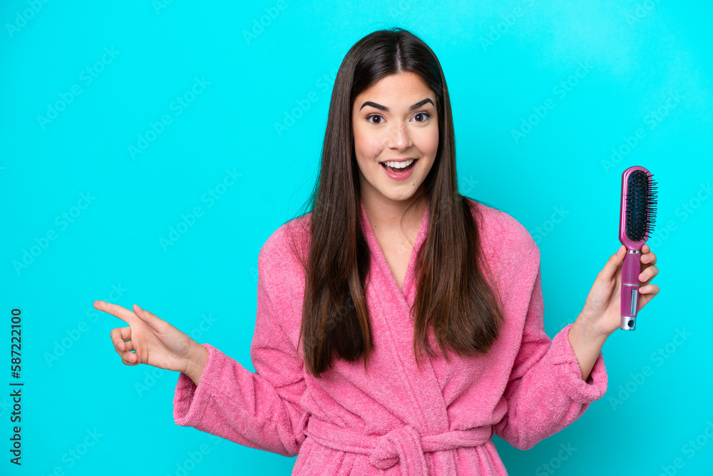 Young Brazilian woman holding hairbrush isolated on blue background surprised and pointing finger to the side Young Brazilian woman holding hairbrush isolated on blue background surprised and pointing finger to the side