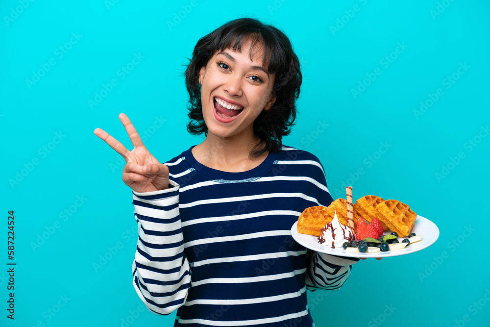 Young Argentinian woman holding waffles isolated on blue background smiling and showing victory sign