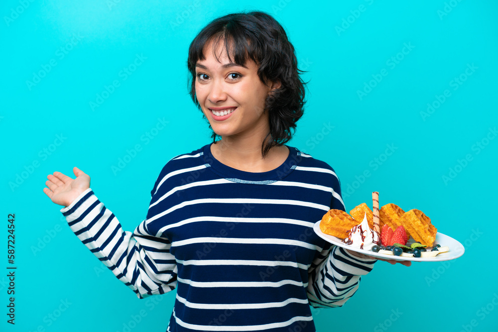 Young Argentinian woman holding waffles isolated on blue background extending hands to the side for inviting to come Young Argentinian woman holding waffles isolated on blue background extending hands to the side for inviting to come