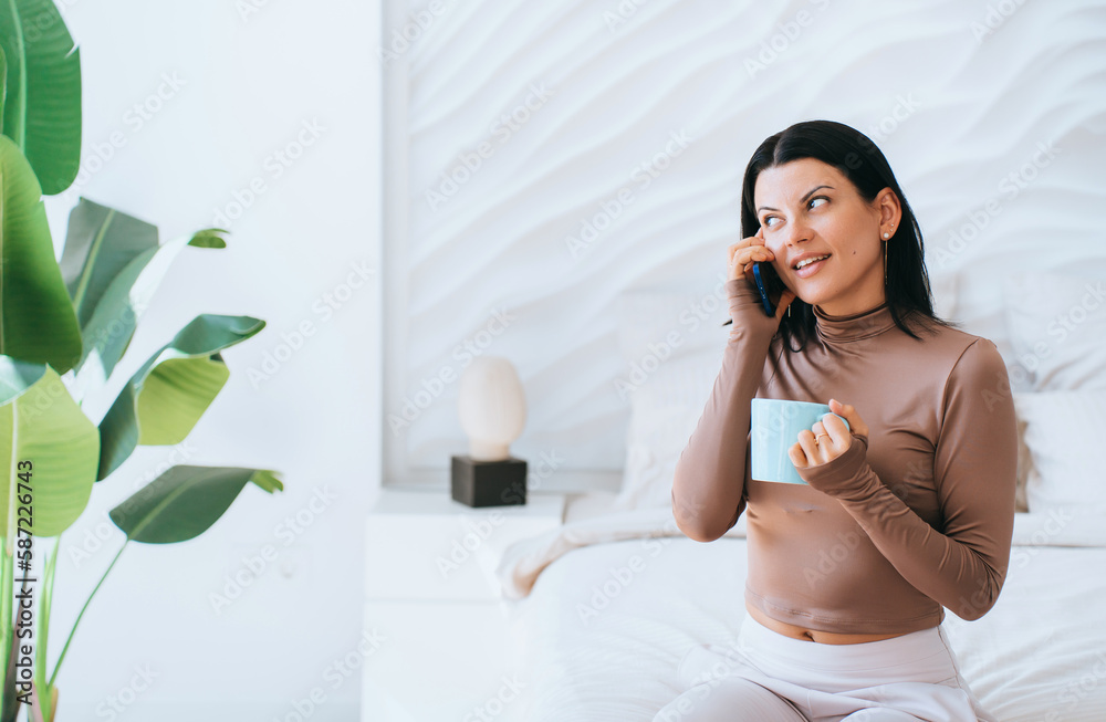 Happy brunette young woman in beige sweatshirt talks by phone, smiles holds cup of coffee at home, great news. Excited business woman received good information, successful deal. Amazement. Happy brunette young woman in beige sweatshirt talks by phone, smiles holds cup of coffee at home, great news. Excited business woman received good information, successful deal. Amazement.