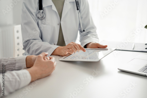 Doctor and patient sitting at the desk in clinic office. The focus is on female physician s hands using tablet computer  close up. Perfect medical service and medicine concept