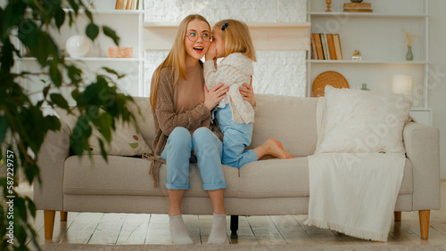 Caucasian family at living room. Woman mother babysitter listen secret from little daughter kid baby child girl whisper talking gossip in mommy ear share secrecy information rumor sitting at couch photo