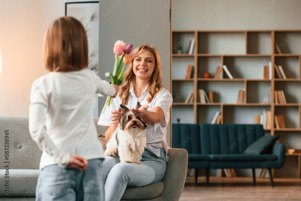 Girl is giving flowers for a woman, holidays. Mother with her little daughter is together in domestic room Girl is giving flowers for a woman, holidays. Mother with her little daughter is together in domestic room