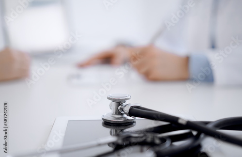 Stethoscope lying on the tablet computer in front of a doctor and patient sitting opposite each other at the background . Medicine, healthcare concept