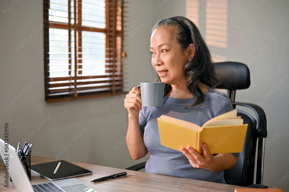 Happy and relaxed Asian-aged businesswoman is reading a book, sipping her morning coffee Happy and relaxed Asian-aged businesswoman is reading a book, sipping her morning coffee