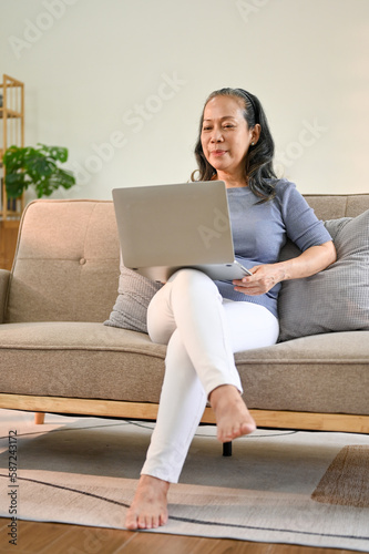 Happy Asian-aged retired woman using her laptop on sofa in her living room.