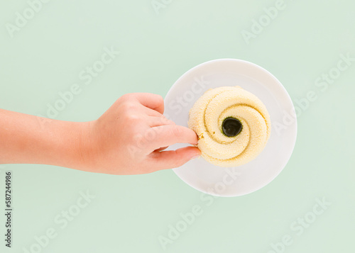 Child's hand dipps fingers into cake on round white plate on isolated pastel green background. Minimal flat lay. Creative concept of sugar or sweet food addiction. Yummy nut dessert. Top view.