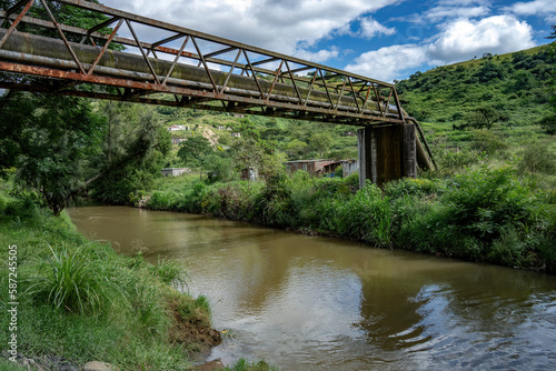 A rusty bridge carrying pipelines is built over a river in a rural area.