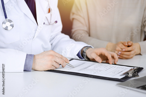 Unknown male doctor and patient woman discussing something while sittingin a darkened clinic and using clipboard, glare of light on the background