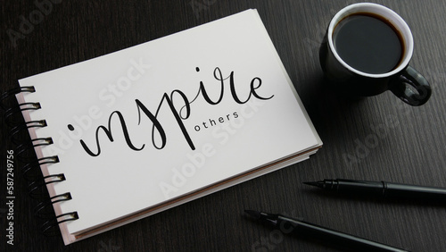 INSPIRE OTHERS black hand lettering in notebook with cup of espresso and pens on black wooden desk