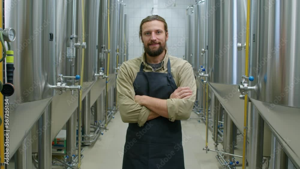 Medium portrait shot of cheerful bearded young Caucasian male technician in apron standing at his workplace in craft brewery, crossing arms on chest, looking at camera and smiling confidently Medium portrait shot of cheerful bearded young Caucasian male technician in apron standing at his workplace in craft brewery, crossing arms on chest, looking at camera and smiling confidently