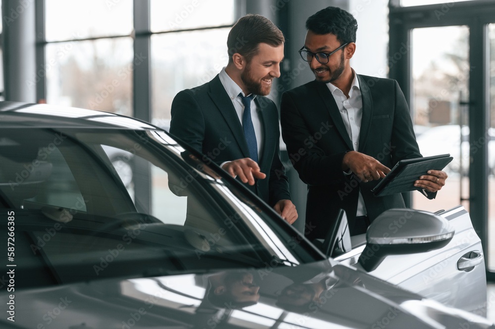 New plan. Standing near automobile with graphic tablet. Two businessmen are working together in the car showroom New plan. Standing near automobile with graphic tablet. Two businessmen are working together in the car showroom