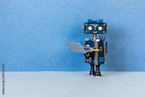 Toy robot bot holds an open compact silver laptop. Modern artificial intelligence machine learning technologies. Copy space on blue background