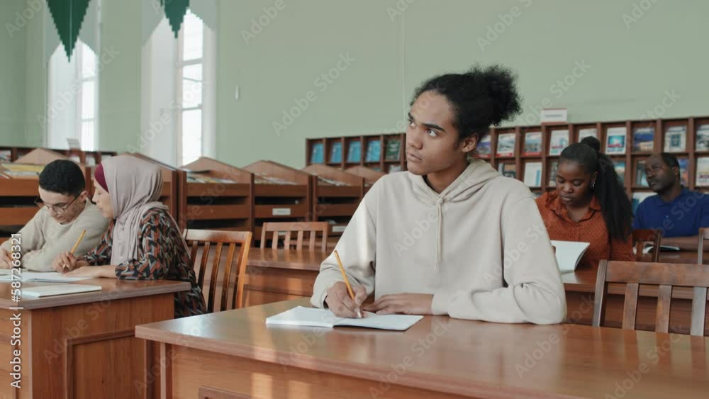 Young Black man and his multi-ethnic classmates sitting at desks making notes during lecture Young Black man and his multi-ethnic classmates sitting at desks making notes during lecture