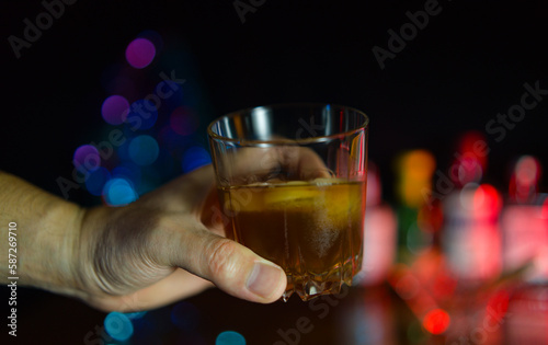 A man's hand clutching a glass with a strong drink against a background of colorful lights of festive atmosphere.