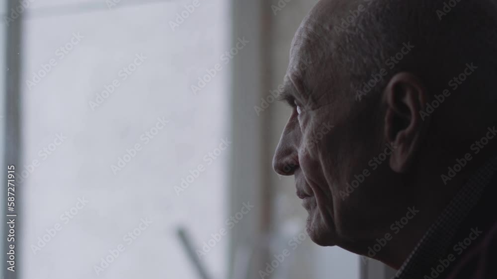 Elderly person looks out window at home and enjoys spending quiet life. Portrait of lonely old wrinkled man thinking about future life