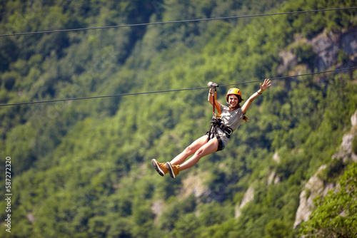 Happy young cheerful female tourist wearing casual clothes riding on zipline in forest.  Zipline trip selective focus against blurred background of forest. Holiday, adventure, extreme sport concept.