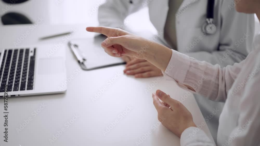 Doctor and patient sitting and discussing something near each other at the white desk in clinic. Unknown female physician pointing into laptop computer screen. Medicine concept Doctor and patient sitting and discussing something near each other at the white desk in clinic. Unknown female physician pointing into laptop computer screen. Medicine concept