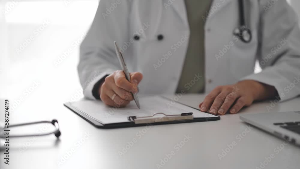 Unknown doctor woman sitting and writing notes at the white desk in clinic or hospital office, close up. Medicine concept