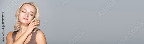 Blonde woman with problem skin touching cheek while standing isolated on grey, banner.