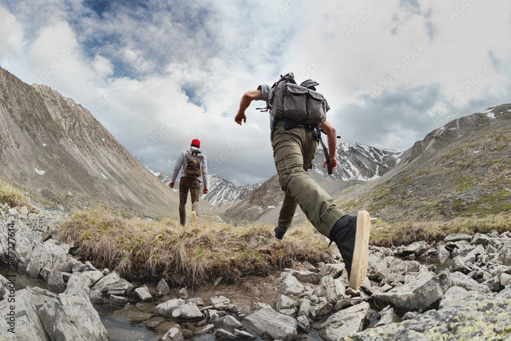 Two young hikers walks with light backpacks in mountains. Tourist jumps across the obstacle - obrazy, fototapety, plakaty 