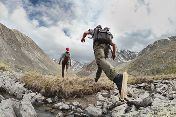 Two young hikers walks with light backpacks in mountains. Tourist jumps across the obstacle