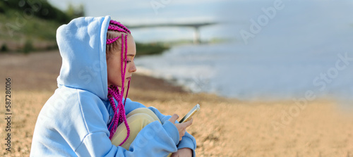Portrait of a caucasian teenage girl with pink braids using smartphone on the river bank.Technology,summer concept.Generation Z style.Copy space for text.