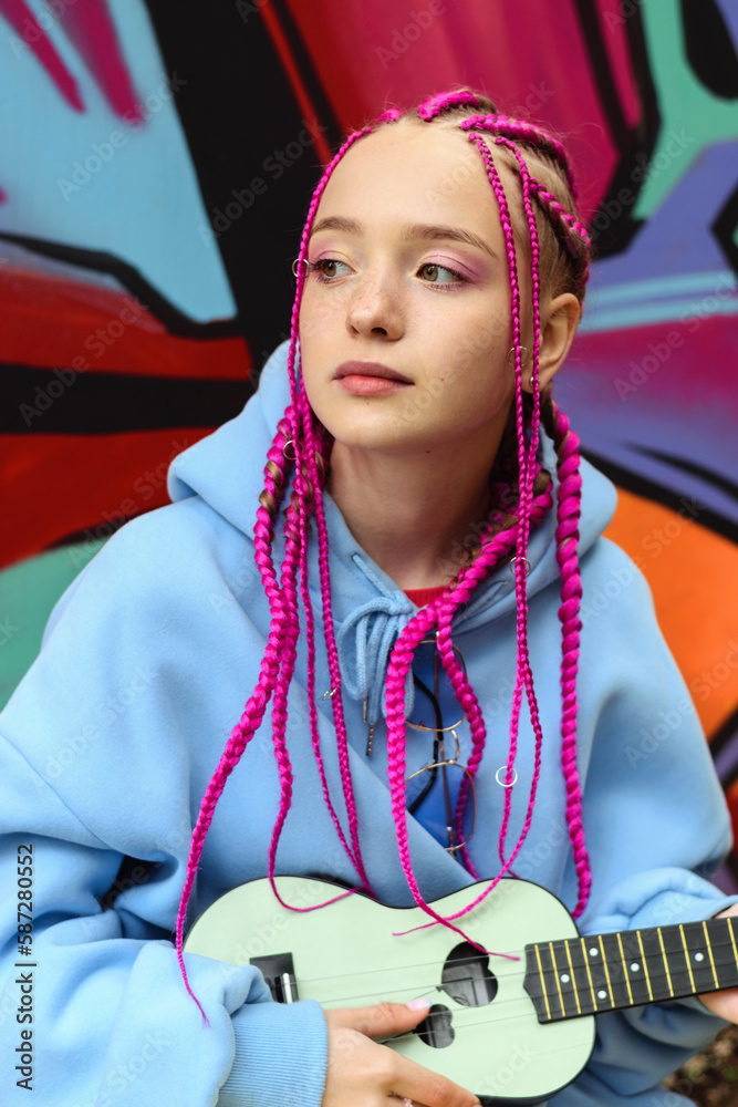 Φωτογραφία Caucasian teenage hipster girl with pink braids plays the ukulele against the background of a multicolored street wall