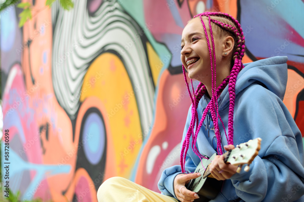 Smiling caucasian teenage hipster girl with pink braids plays the ukulele against the background of a multicolored street wall.Summer concept.Generation Z style.