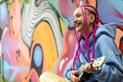 Obraz na płótnie Smiling caucasian teenage hipster girl with pink braids plays the ukulele against the background of a multicolored street wall.Summer concept.Generation Z style. w salonie