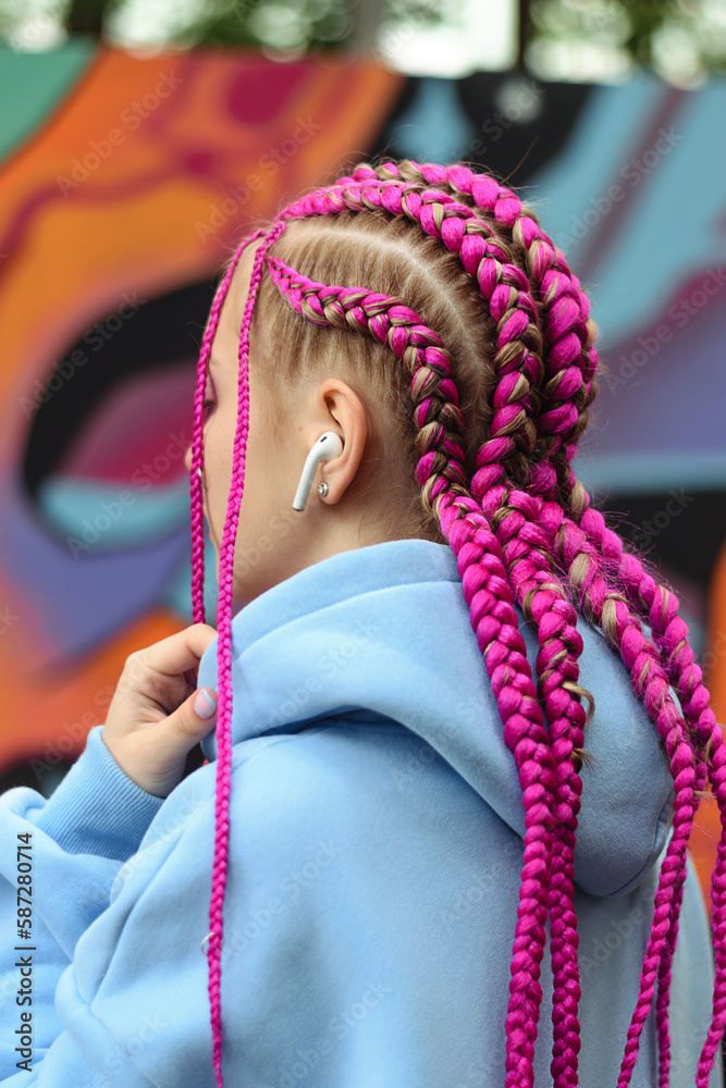 Caucasian teenage girl with pink braids using wireless headphones against the background of a multicolored street wall.Generation Z style,creativity.