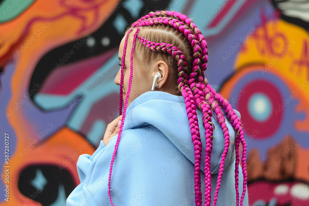 Caucasian teenage girl with pink braids using wireless headphones against the background of a multicolored street wall.Generation Z style,creativity.