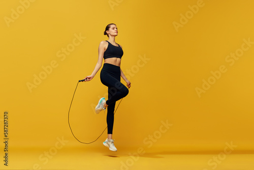 Sportive young girl with fit body training, jumping over jumping rope against yellow studio background. Cardio workout. Concept of sport, healthy and active lifestyle, beauty, fitness © master1305