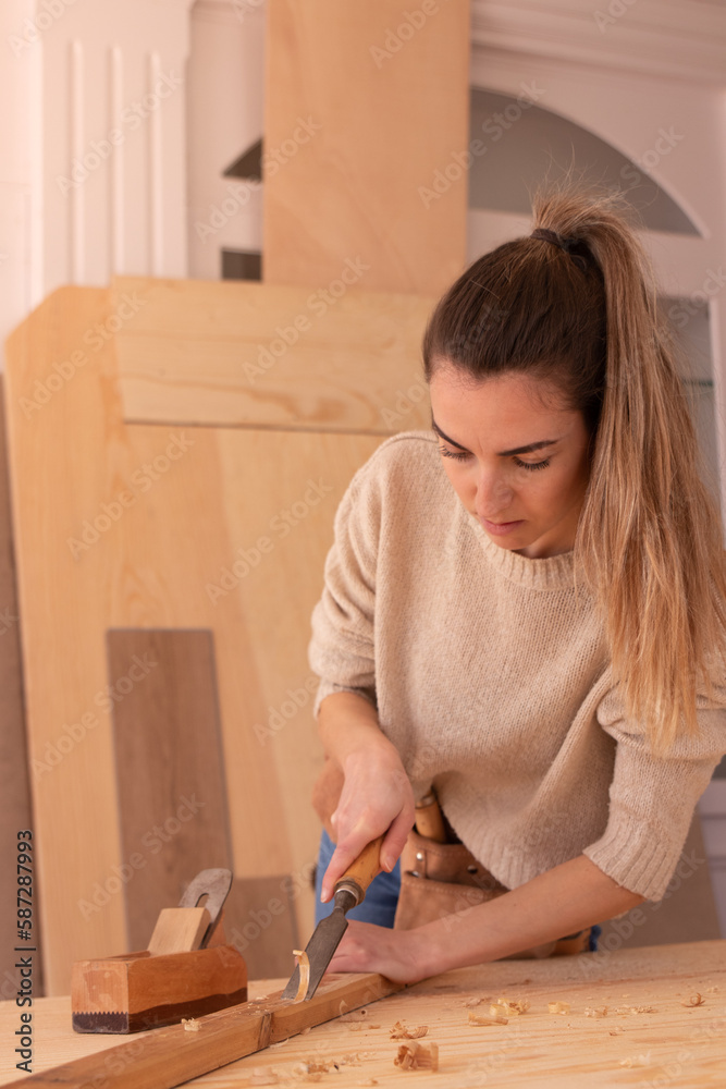 Handy woman gouging wood with chisel. Focused female woodworker in apron carving wood with rasp near jack plane on workbench Handy woman gouging wood with chisel. Focused female woodworker in apron carving wood with rasp near jack plane on workbench
