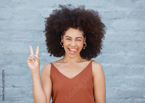 Black woman, portrait and tongue out with peace sign and afro against a gray wall background. Happy and goofy African female face smile showing peaceful hand emoji, sign and funny or silly expression