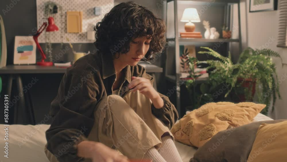 Young girl with curly short hair checking her phone, then putting it away, looking depressed and disappointed, touching her neck and face, sitting on unmade bed, desk, lamps and plant in background Young girl with curly short hair checking her phone, then putting it away, looking depressed and disappointed, touching her neck and face, sitting on unmade bed, desk, lamps and plant in background