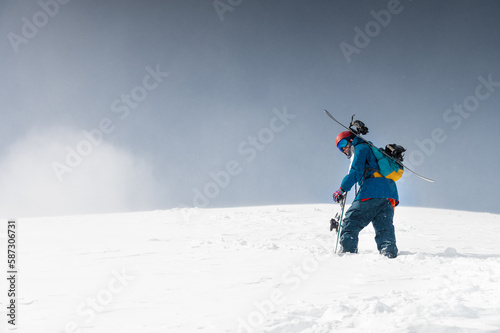 man with ski equipment and a snowboard climbs a snowy mountain. Blue sky and snowy mountain in the background.