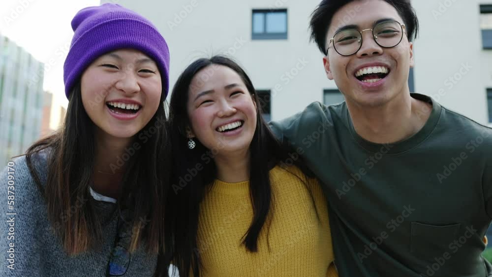 United young teenage asian friends hugging each other laughing outdoors. Three colleague ethnic student having fun together in city street.