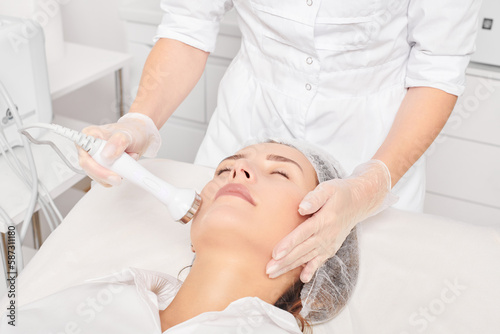 Fotografie Beautician makes ultrasound skin tightening for rejuvenation woman face using phonophoresis, anti aging cosmetic procedure with in beauty spa salon