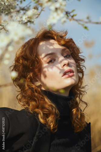 a young, beautiful red-haired woman near a flowering tree poses looking at the camera