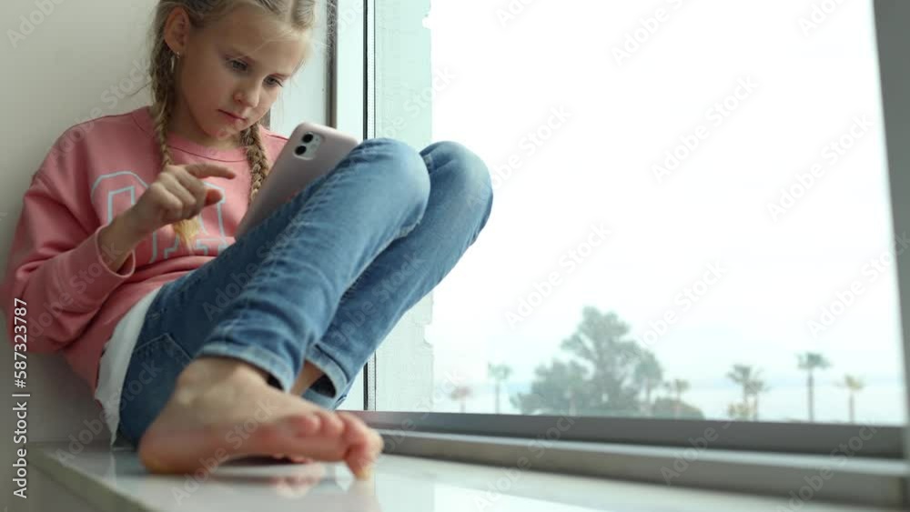 Cute serious little girl sitting barefoot on windowsill and playing in smartphone