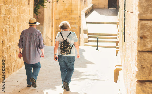 An unrecognizable mature white Caucasian couple of tourists walking through a typical Mediterranean village - wearing backpack and straw panama hat