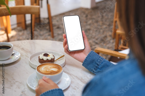 Mockup image of a woman holding and using mobile phone with blank desktop screen while drinking coffee in cafe