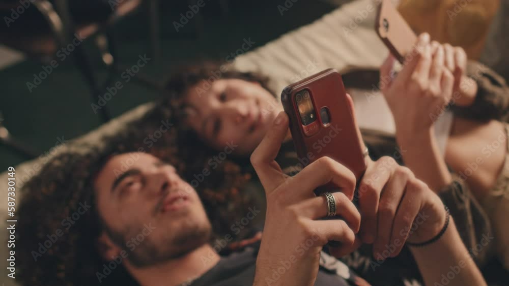 Young couple lying in bed with their phones, looking at each other screens, talking, smiling. Sunlight, high angle view, blurred Young couple lying in bed with their phones, looking at each other screens, talking, smiling. Sunlight, high angle view, blurred